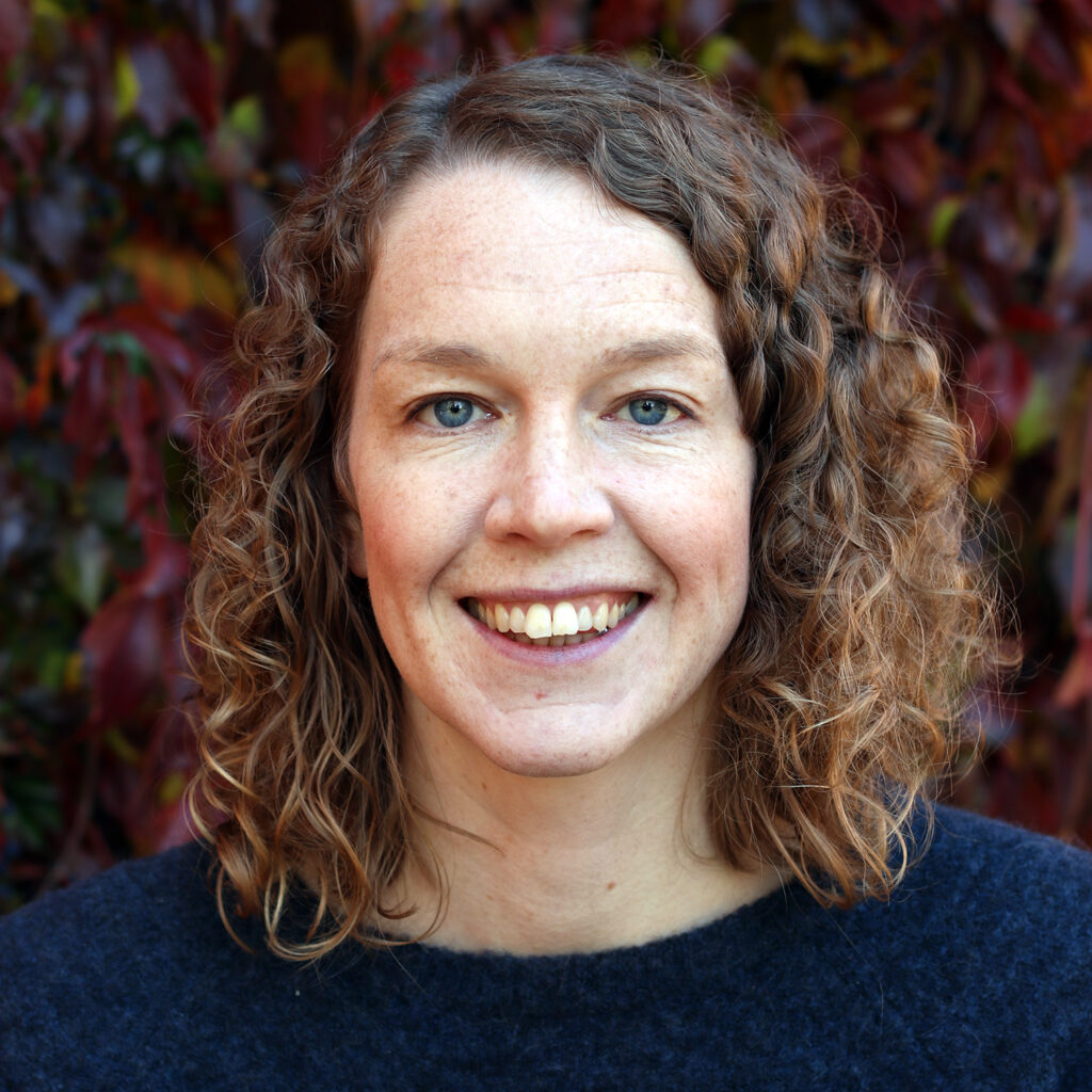 Matilda Morin smiling in an outdoor portrait with curly hair and autumn-colored foliage in the background.