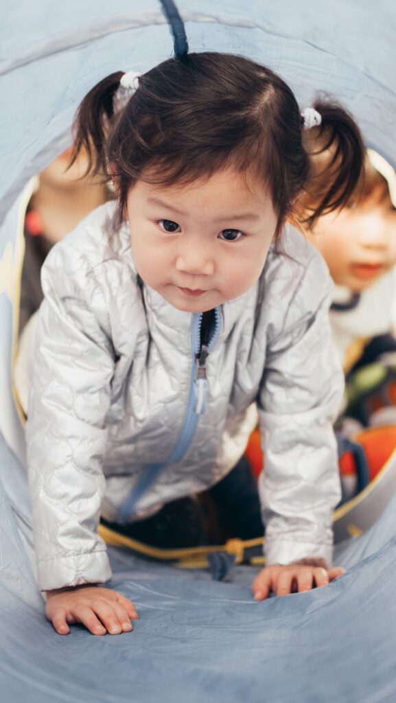 Young child crawling through a fabric play tunnel, with another child behind them.